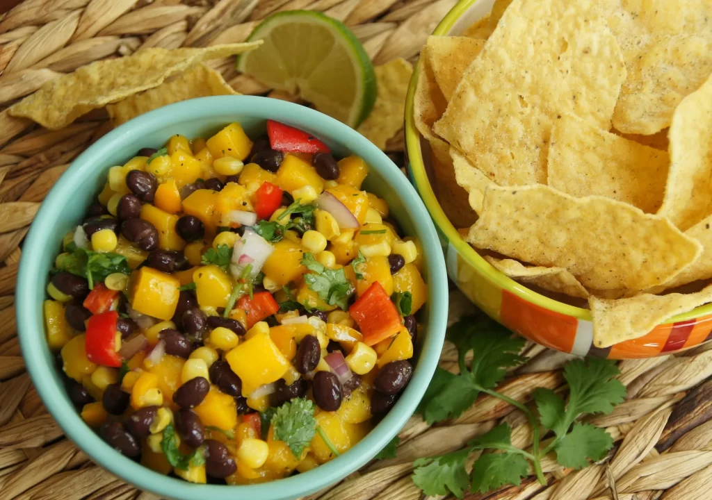 A woven placemat with a bowl filled with mango, black beans, red pepper, onion and cilantro. Another bowl is filled with chips. There is a lime wedge and cilantro.