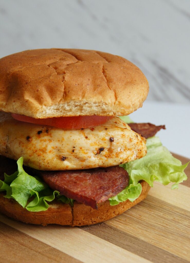 A wooden board with a chicken sandwich. Topped with lettuce, bacon and tomato.