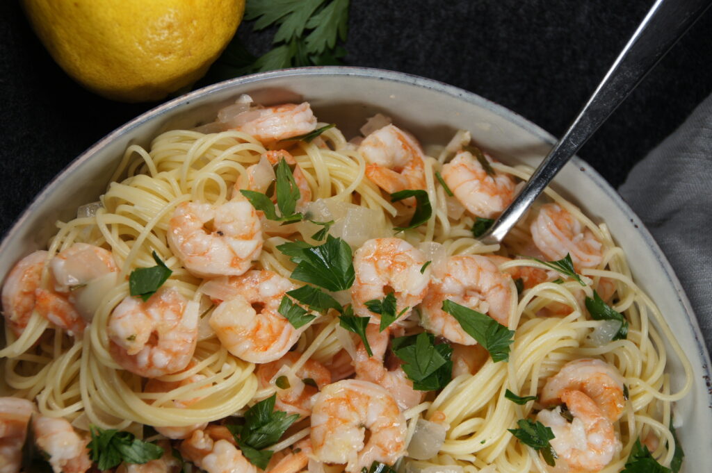 A bowl with noodles, shrimp scampi, onion and parsley. A lemon is on the side.