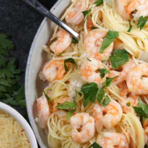 A bowl with noodles, shrimp scampi, onion and parsley. And a small bowl with shredded parmesan.