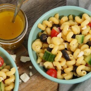 Two bowls filled with cavatappi pasta, tomatoes, black olives, cucumbers, parsley and feta cheese. A greek vinaigrette sits on the side.
