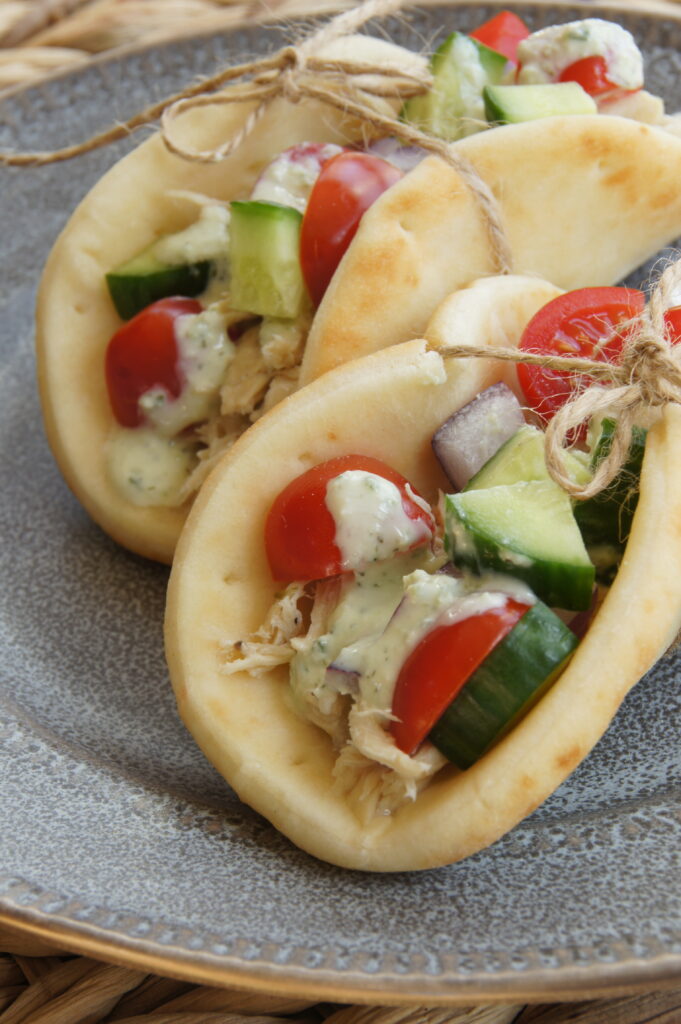 A plate with two gyros tied together. The gyros are filled with shredded chicken and chunks of tomatoes and cucumbers and slices of red onion. All topped with a tzatziki sauce.