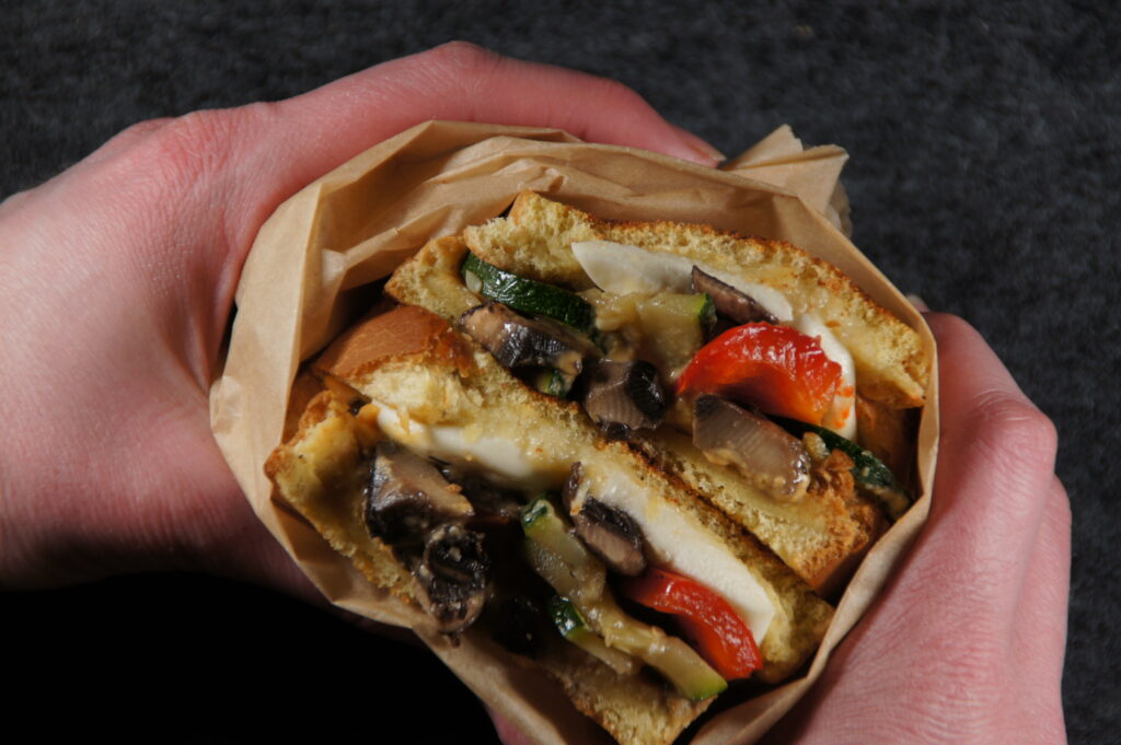Two hands holding a panini that is filled with lots of vegetables and cheese and wrapped in brown paper.