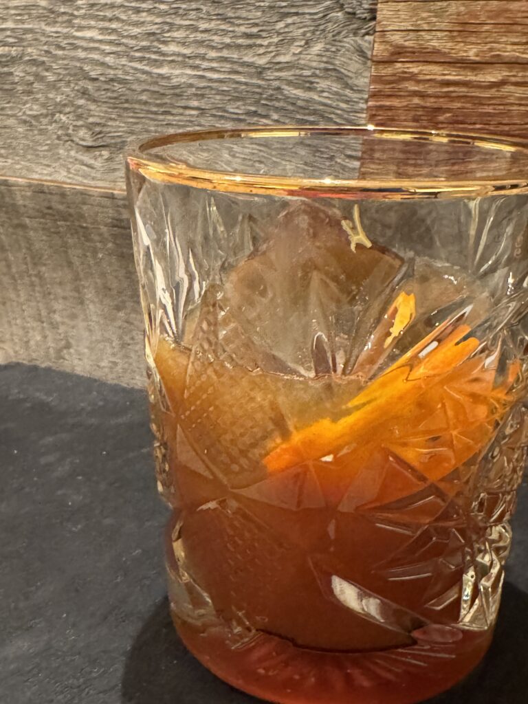 A lowball glass filled with a large ice cube and cinnamon cardamom simple syrup. Topped with bourbon and orange peel. Cinnamon sticks on the side.