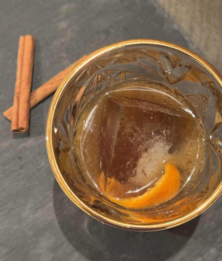 A lowball glass filled with a large ice cube and cinnamon cardamom simple syrup. Topped with bourbon and orange peel. Cinnamon sticks on the side.