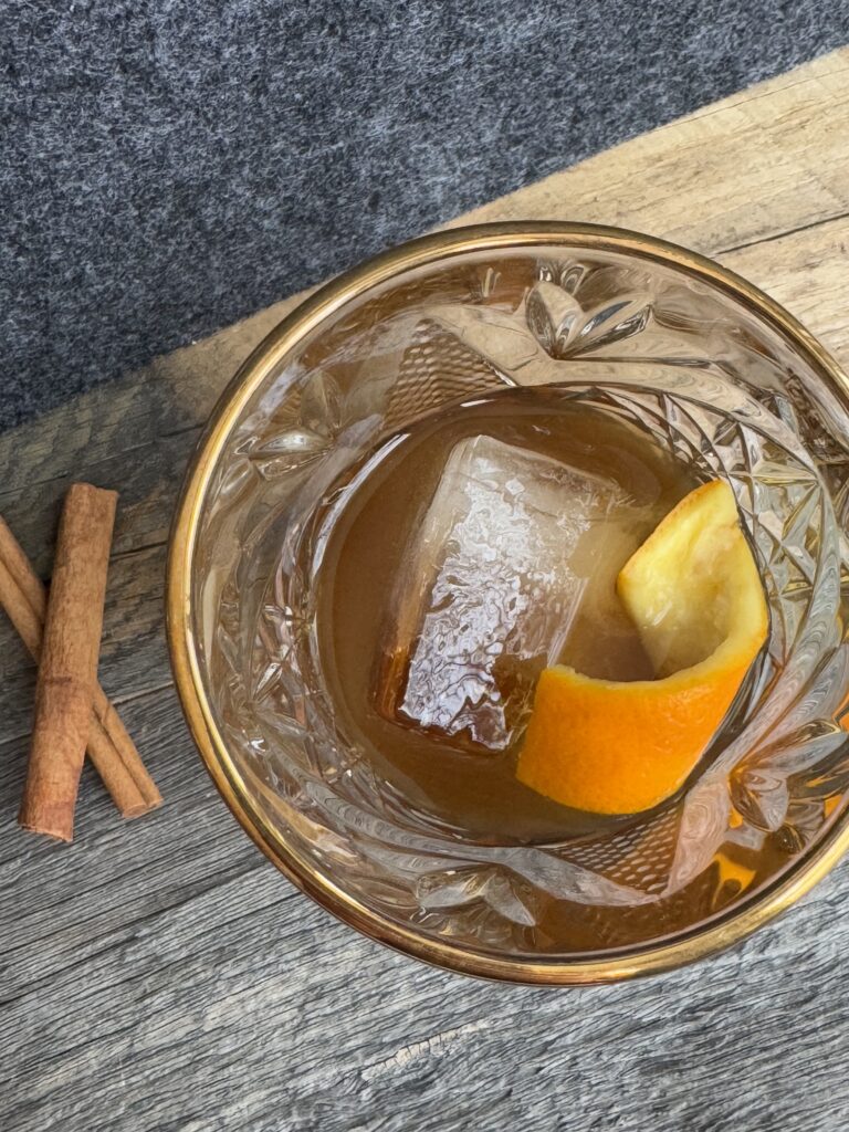 A lowball glass filled with a large ice cube and cinnamon cardamom simple syrup. Topped with bourbon and orange peel. Cinnamon sticks on the side.