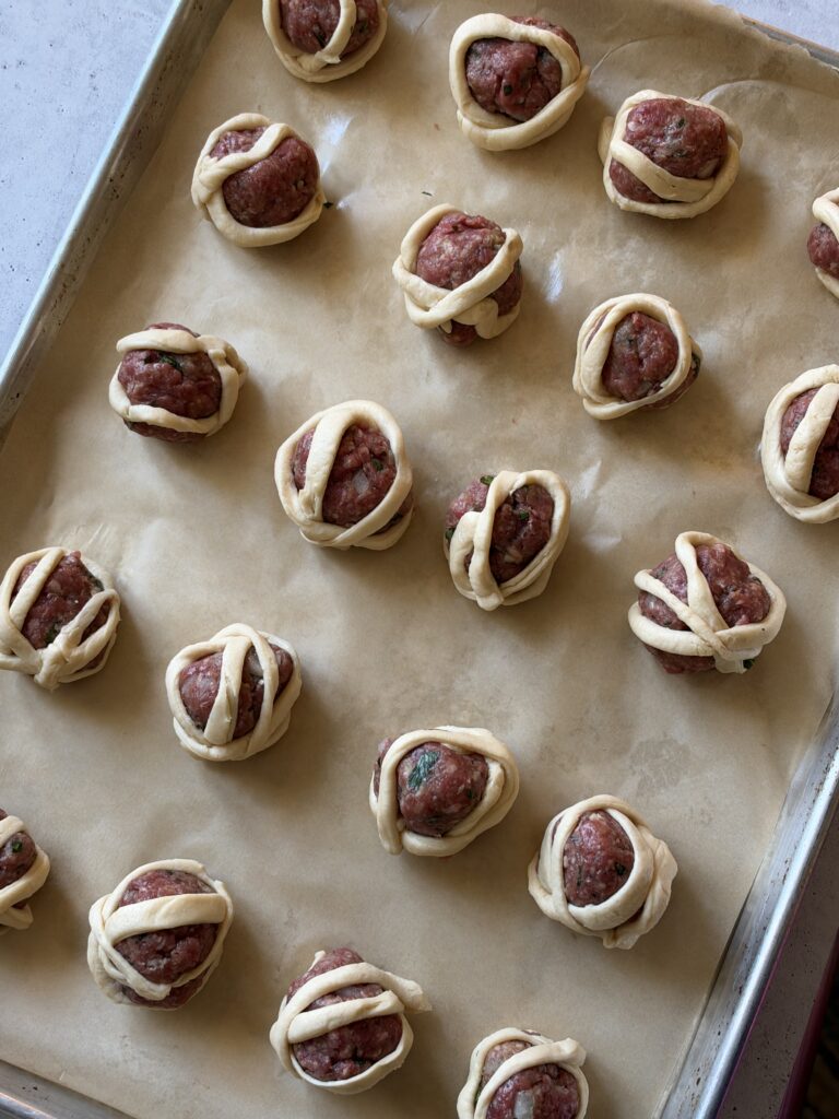 A sheet pan with small meatballs and thing strips of flaky dough wrapped around them to look like mummies.