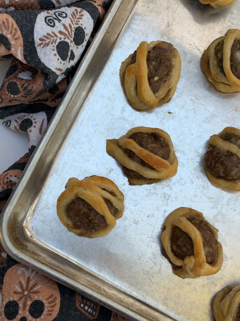 A silver pan is filled with meatballs wrapped in flaky puff pastry to look like little mummies.
