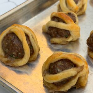 A silver pan is filled with meatballs wrapped in flaky puff pastry to look like little mummies.