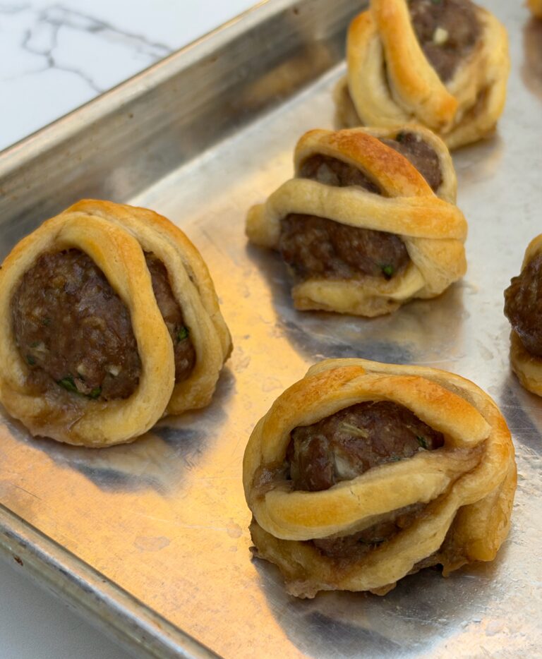 A silver pan is filled with meatballs wrapped in flaky puff pastry to look like little mummies.