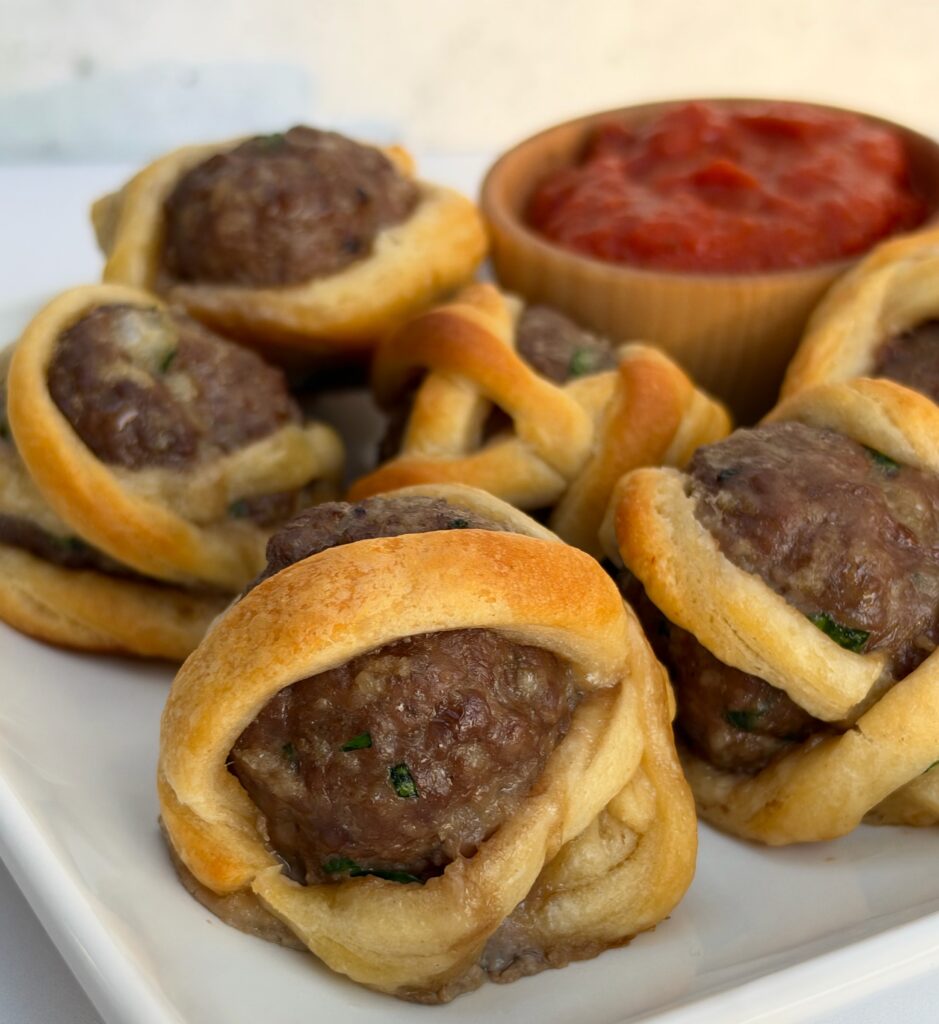A white plate has meatballs on it that are wrapped in strips of dough to look like mummies. A bowl of marinara sits on the side for dipping.