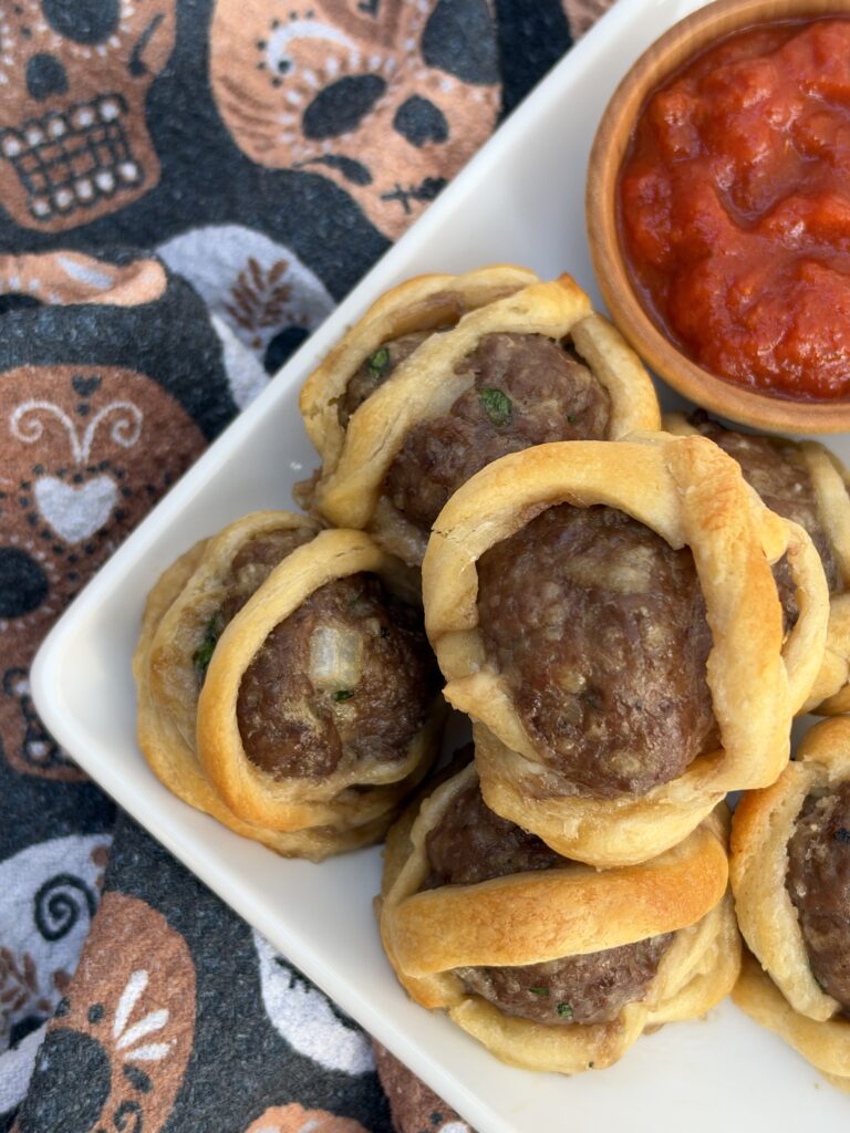 A plate filled with meatballs that are wrapped with dough. There is a bowl on the side with marinara and a skeleton towel in the background.