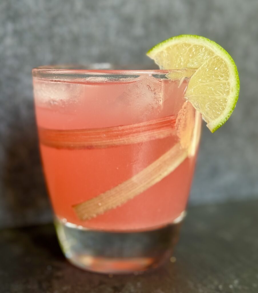A glass filled with ice and some gin. Topped with tonic water and rhubarb simple syrup. A wedge of lime is on the side.