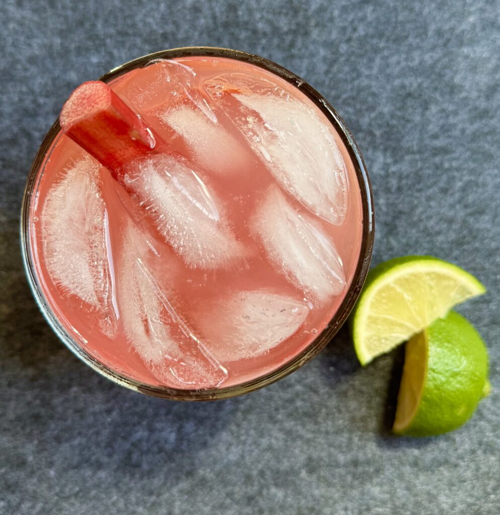 A glass filled with ice and some gin. Topped with tonic water and rhubarb simple syrup. A wedge of lime is on the side.