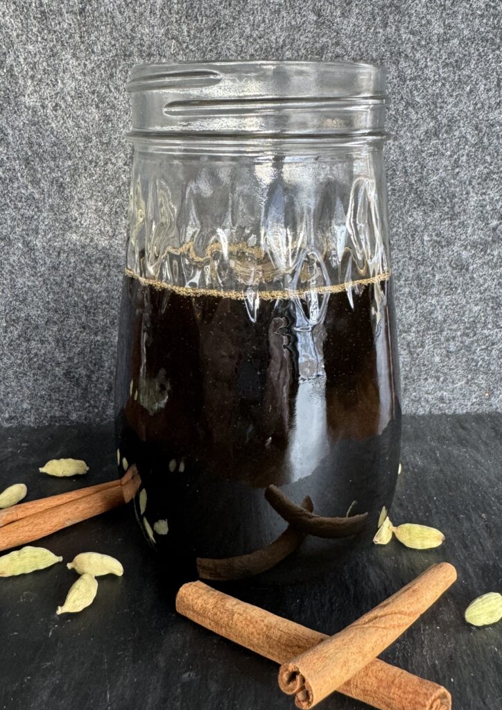 A glass jar filled with a cinnamon cardamom simple syrup with cinnamon sticks and cardamom pods in the foreground.