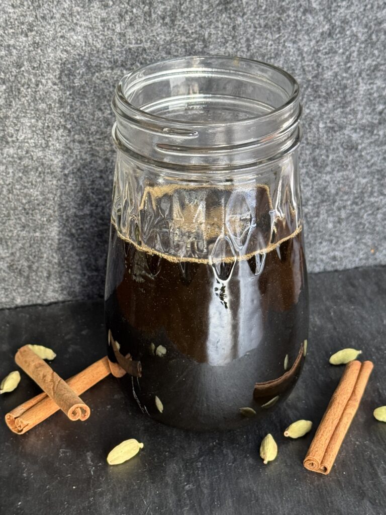 A glass jar filled with a cinnamon cardamom simple syrup with cinnamon sticks and cardamom pods in the foreground.