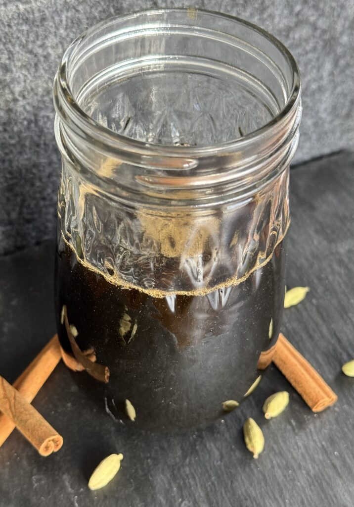 A glass jar filled with a cinnamon cardamom simple syrup with cinnamon sticks and cardamom pods in the foreground.