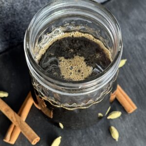 A glass jar filled with a cinnamon cardamom simple syrup with cinnamon sticks and cardamom pods in the foreground.