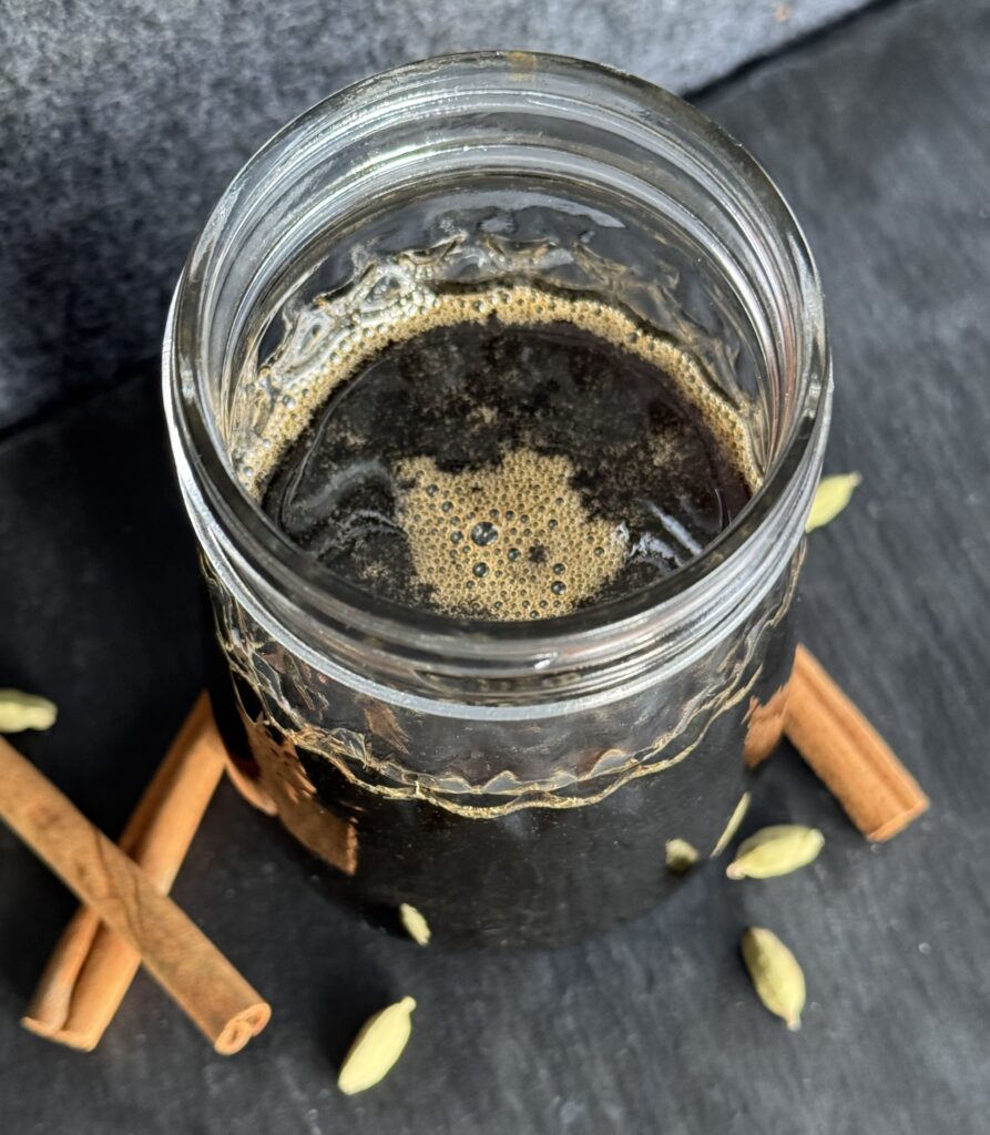 A glass jar filled with a cinnamon cardamom simple syrup with cinnamon sticks and cardamom pods in the foreground.