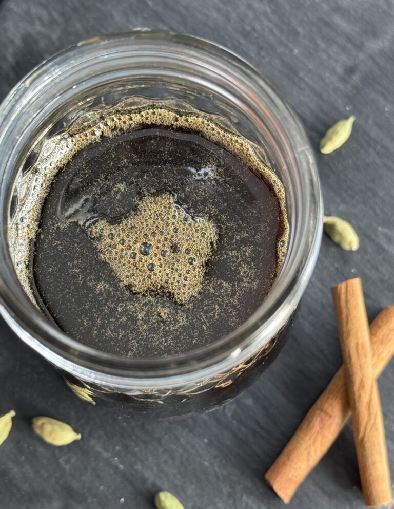 A glass jar filled with a cinnamon cardamom simple syrup with cinnamon sticks and cardamom pods in the foreground.